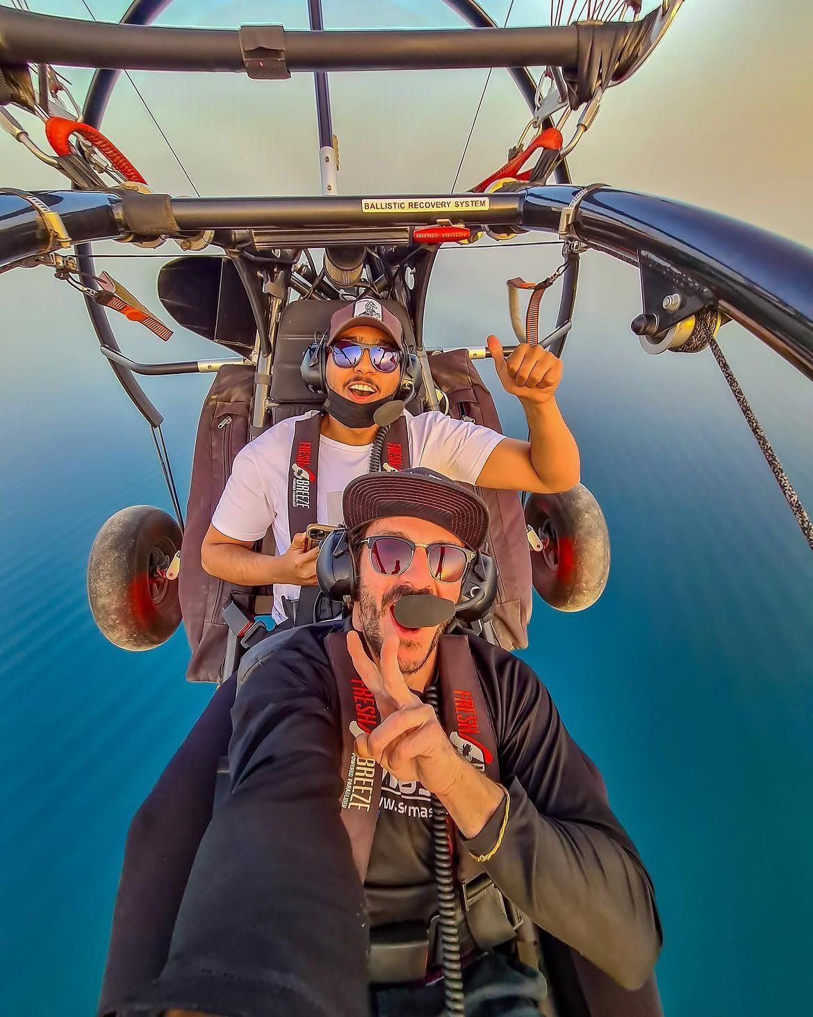 Javi flying a paratrike with a young passenger, both giving peace signs